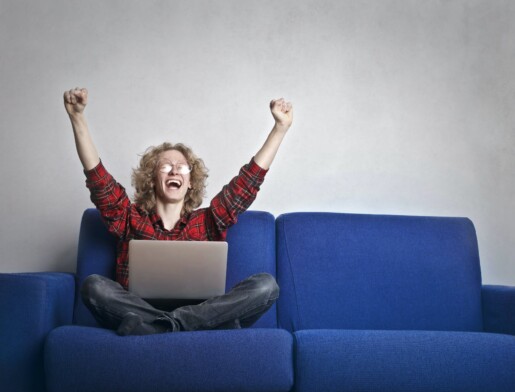 photo of excited person with hands up sitting on a blue sofa while using a laptop